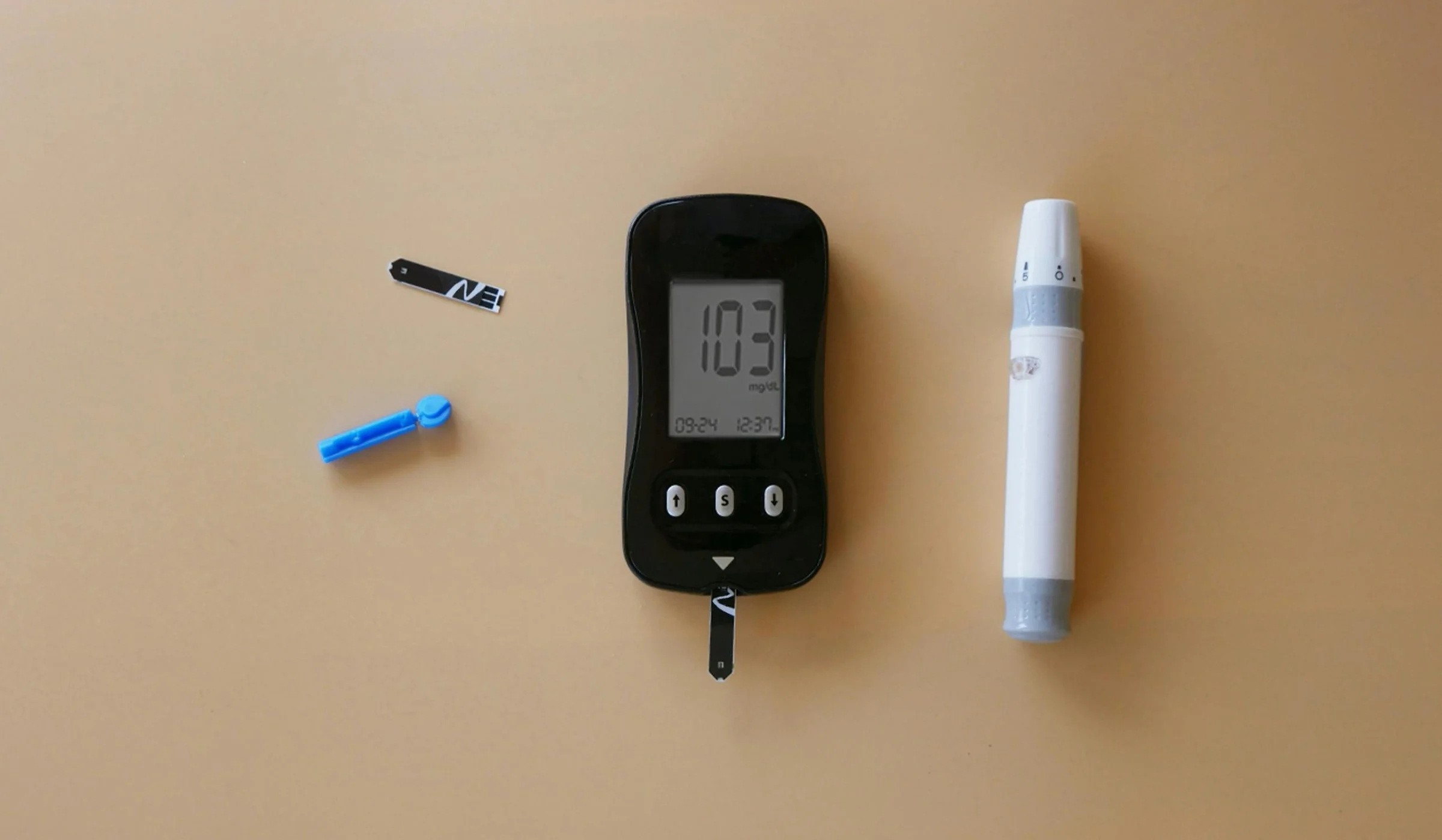 Blood glucose meter reading 103 mg/dL beside a test strip, lancet and lancing device on a tan background
